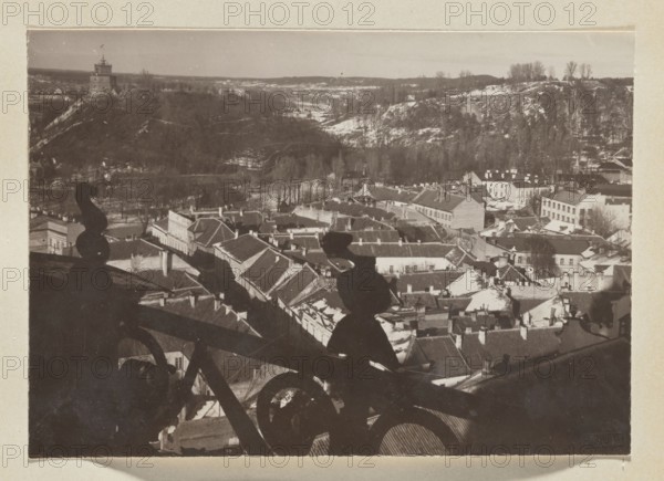 Castle Hill and City Centre; photograph, Jan Bulhak 1876–1950, panorama of Vilnius, 1912; photograph, photographic print: height 11.2 cm, width 16.0 cm; album page: height 31.0 cm, width 23.5 cm, A sepia-toned photograph shows a cityscape from a high vantage point with a foreground of dark ornamental metal railings casting shadows, a middle ground of closely packed rooftops and buildings with varied rooflines and chimneys, and a background of tree-covered hills and a distant stone tower on a hill. The image contains shades of brown, black, tan, and off-white, with lighter areas of snow or pale rooftops contrasting against darker roofs, railings, and tree shadows.