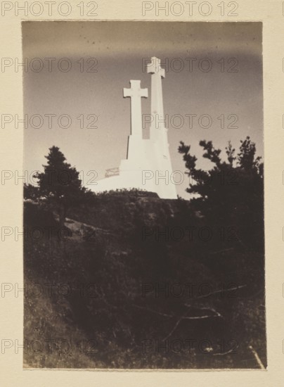 Three Crosses; photograph, volume 1, Bulhak, Jan 1876–1950, Vilnius Lithuania, Wiwulski, Antoni 1877–1919, monuments, Mount Trzykrzyska Vilnius, after 1916, photograph, photographic print: height 15.2 cm, width 10.7 cm, album page: height 31.0 cm, width 23.5 cm, A tall pale monument with two vertical cross-shaped structures rises above a dark hilltop, contrasted against a light sky; the monument appears bright white while the foreground hill and vegetation are deep black and dark brown. A few silhouetted trees with irregular branches flank the monument on either side, showing dark greenish-black tones. The surrounding sky shows gradients of pale beige to muted gray, and the image is framed by a textured beige border.