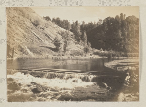 Wilejka in the Bernardine Garden; photograph, 1912; photographer Jan Bulhak 1876–1950; place of creation: Vilnius, Bernardine Garden; photographic print: 11.5 × 16.0 cm; album page: 31.0 × 23.5 cm., A sepia-toned photograph shows a river flowing over a low waterfall with white, foamy water cascading over a smooth ledge and swirling around dark rocks in the foreground; a log lies along the right riverbank. Behind the falls the river widens into a calm stretch reflecting light, bordered by grassy banks and a gently sloping hillside on the left that is dotted with shrubs and small trees. A group of taller trees forms a dense, darker stand on the right side of the image, creating contrast with the lighter hillside. The overall colors visible are shades of brown, beige, tan, cream, and darker brown.