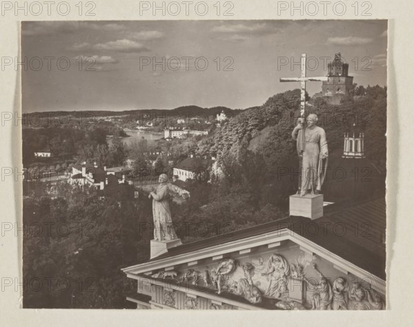 The Cathedral and Antakalnis; photograph, Jan Bulhak 1876–1950, panorama of Vilnius showing the cathedral and Castle Hill, 1912; photographic print: 11.5 × 16.0 cm; album page: 31.0 × 23.5 cm., The image shows a sepia-toned scene with a rooftop foreground featuring sculpted figures and reliefs in light beige tones, a large statue of a person holding a tall cross in similar pale stone, and another statue in prayer posture. Behind the rooftop is a dark roof dome and a lighter stone pedestal. The middle ground contains a hillside covered in dense trees in shades of dark green and brown, and a cluster of buildings with light-colored facades and darker roofs. The background reveals a river or body of water bordered by more tree-covered land and distant low hills under a cloudy sky in muted gray and beige tones.