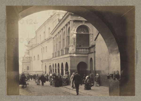 The Gate of Dawn; photograph, 1912, Vilnius; photographer Jan Bulhak 1876–1950; photographic print: 11.0 × 15.7 cm; album page: 31.0 × 23.5 cm., The image shows a sepia-toned street scene framed by a large dark arch in the foreground, with a lighter stone building featuring a row of arches and a columned upper gallery occupying the middle ground; several people are gathered along the sidewalk near the arches, many wearing long garments and head coverings, and a few figures are walking along the street; a cart with a covered load is visible to the left, and a group of people sit or stand near the base of the building to the right; the visible colors are shades of brown, beige, black, and gray.