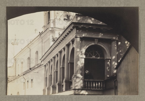 The Gate of Dawn; photograph, 1912, Vilnius; photographer Jan Bulhak 1876–1950; photographic print: 11.0 × 16.5 cm; album page: 31.0 × 23.5 cm., The image shows an exterior view of a building with a colonnaded balcony and arched openings; the facade displays columns, balustrades, and an arched window with a fanlight set above a dark opening where a person wearing a hat is partially visible; light and shadow create dappled patterns on the walls and columns; colors visible include shades of beige, cream, gray, and dark brown/black.