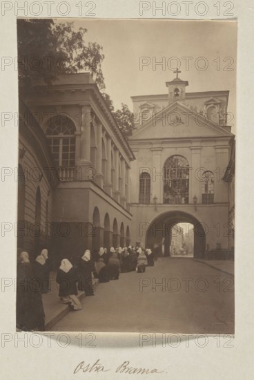The Gate of Dawn; photograph, 1912; Bulhak, Jan 1876–1950; Vilnius Lithuania; photograph, photographic print: height 15.8 cm, width 11.0 cm; album page: height 31.0 cm, width 23.5 cm, The image shows a narrow street flanked by a large classical building with columns and arches on the left and a façade with a triangular pediment and a central arched passage on the right; a group of people wearing dark clothing and white head coverings are kneeling or sitting along the left side of the street; the scene includes architectural details such as tall arched windows, balustrades, and a small cross atop the central façade; colors visible are sepia tones, brown, beige, off-white, and dark gray.