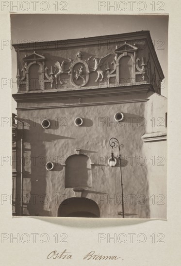 The Gate of Dawn; photograph, 1912. Photograph in the album Vilnius, volume 1, Jan Bulhak 1876–1950. Vilnius. Photographic print: 13.6 × 10.5 cm; album page: 31.0 × 23.5 cm., A vertical sepia-toned photograph shows the façade of an ornate building with a textured light-colored wall and a decorative upper frieze featuring sculpted figures and architectural niches. The top section has sculptural reliefs including winged creatures and framed medallion shapes, with ornamental columns and arch-like elements in pale tones. Below, the plain wall contains a group of circular openings that cast darker circular shadows, and a recessed arched niche above a larger dark arched doorway. A tall street lamp with a curved ornate arm and a globe-shaped lantern stands in front of the wall, casting a shadow. The image border is pale and the overall palette consists of brown, beige, cream, and darker brown shadows.
