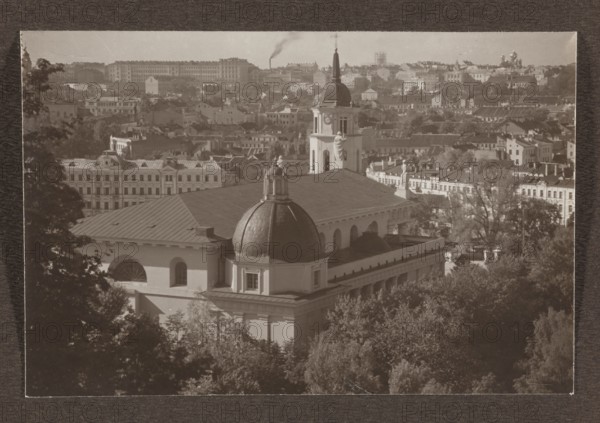 Cathedral. General view; photograph titled Vilnius, volume 1, Bulhak, Jan 1876–1950, panorama of Vilnius, 1912; photograph, photographic print: height 10.5 cm, width 15.7 cm; album page: height 31.0 cm, width 23.5 cm., The image shows an aerial view of a cityscape with a large classical building in the foreground featuring a prominent dome and a tall tower topped with a spire, flanked by statues on the roofline; the building has a light exterior and a darker domed roof. Surrounding the main building are trees and foliage in the lower part of the image and a dense cluster of rooftops and multi-story buildings extending into the background. The skyline includes a variety of roof shapes and architectural details, and there is a vertical plume of smoke rising in the distance. The overall image tones are sepia, showing shades of brown, beige, cream, and darker brown.