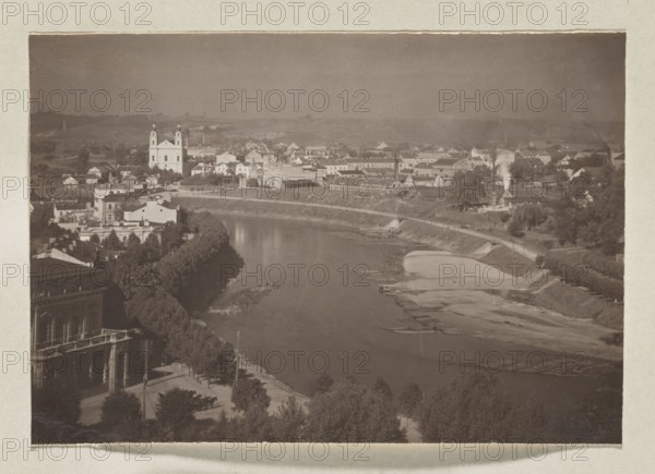 Wilia towards the Green Bridge; photograph, Wilno album, volume 1; Jan Bulhak 1876–1950; Vilnius panoramas; Wilia river, panorama of Vilnius; 1912; photograph, photographic print: 11.0 × 16.0 cm; album page: 31.0 × 23.5 cm, The image shows a riverside urban scene with a broad river curving through the center, bordered by tree-lined embankments and sandy exposed banks. Along the near bank there is a promenade area with a few trees and a large building with arches and a balcony. Across the river a group of buildings rises on gently rolling land, including a prominent twin-towered church with a light facade and several clustered structures of varied shapes and rooflines. There are a few smaller houses, some industrial-looking buildings emitting a plume of smoke, and a mix of open spaces and rows of trees. The foreground and riverbanks feature darker greens and browns, the buildings display muted light tones, and the sky appears a soft grayish tone.