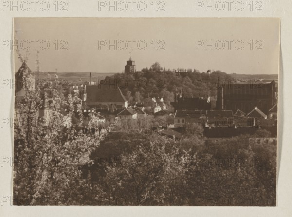 Castle Hill; photograph, Bulhak, Jan 1876–1950, Vilnius, 1912; panorama of Vilnius, Castle Hill; photograph, photographic print: height 11.5 cm, width 16.3 cm; album page: height 31.0 cm, width 23.5 cm, A sepia-toned cityscape showing a foreground of dense trees and blossoming branches in lighter tones, a midground of clustered rooftops and buildings in darker browns and muted beige, and a background of a wooded hill topped by a prominent tower structure; the sky above is pale beige and the overall image contains shades of brown, sepia, beige, and dark brown.