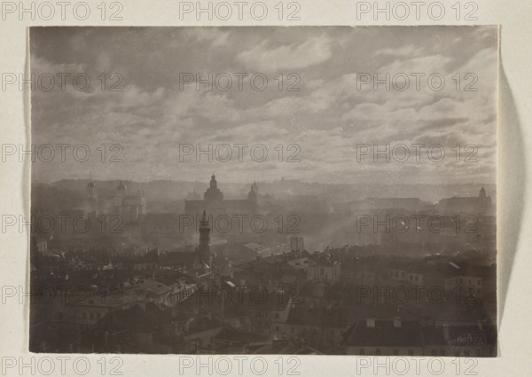 General view from the Stojanska bell tower; photograph, 1912; photographer Jan Bulhak 1876–1950; city panorama of Vilnius; photographic print: 11.0 × 15.7 cm; album page: 31.0 × 23.5 cm., A hazy cityscape fills the image, with a skyline of rooftops and spires receding into mist under a broad, cloud-filled sky. The foreground shows a dense cluster of buildings with pitched and flat roofs in darker shades, while a pointed tower and several domed and cupola-like structures rise above the nearer rooftops. Midground and background layers of buildings fade into lighter, more muted tones through the haze, creating a sense of depth. The sky occupies a large portion of the frame with textured, streaky clouds. Visible colors include shades of gray, muted brown, and creamy off-white around the image border.