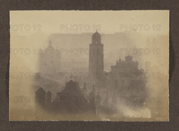 Churches; photograph, Vilnius, volume 1, Jan Bulhak 1876–1950, panoramas, panorama of Vilnius, 1912, Vilnius; photograph, photographic print: height 11.3 cm, width 16.2 cm; album page: height 31.0 cm, width 23.5 cm, A sepia-toned photograph showing a foggy skyline with the silhouettes of a group of ornate buildings and towers. The scene includes a tall central tower flanked by several domed and spired structures, their edges softened by mist. The foreground shows darker silhouettes of rooftops and small towers, while the background fades into lighter, hazy shapes. Visible colors are shades of brown, beige, and gray.