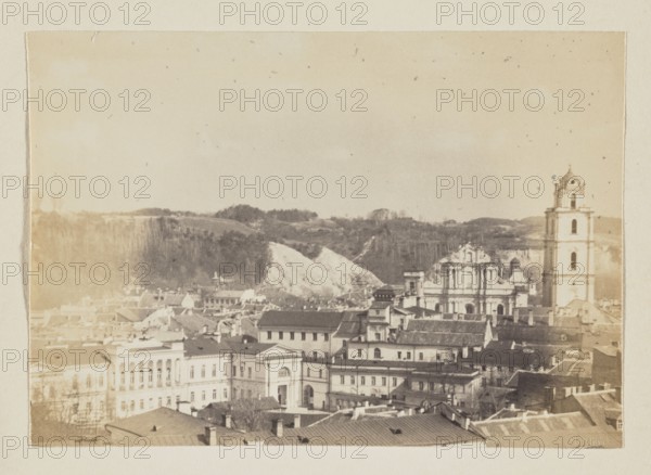 St. John's Church; photograph, 1912, panorama of Vilnius; photographic print: height 11.5 cm, width 16.5 cm; album page: height 31.0 cm, width 23.5 cm; maker: Jan Bulhak 1876–1950; place of origin: Vilnius, Lithuania; subject: panoramas, St. John's Church Vilnius., A sepia-toned panoramic view shows a historic cityscape with a group of closely packed buildings and rooftops in the foreground, including a large ornate church façade and a tall bell tower to the right. Behind the built area there is a hillside with exposed lighter-colored earth and a tree-covered ridge. The sky above is pale with scattered small dark specks. Visible colors are sepia tones, beige, brown, cream, and hints of darker gray.