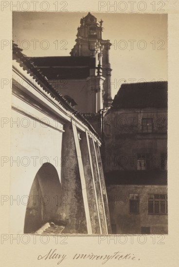 University walls; photograph in the album Vilnius, Bulhak, Jan 1876–1950, St. John's Church Vilnius, 1912, Vilnius, photograph, photographic print: height 16.3 cm, width 11.7 cm, album page: height 31.0 cm, width 23.5 cm, The photo shows an architectural scene with a close view of a light-colored wall and a row of slanted buttresses leading toward a taller ornate tower in the background; a darker building with a tiled roof and several windows sits to the right, and the overall image palette is sepia tones with shades of beige, brown, cream, and dark brown.