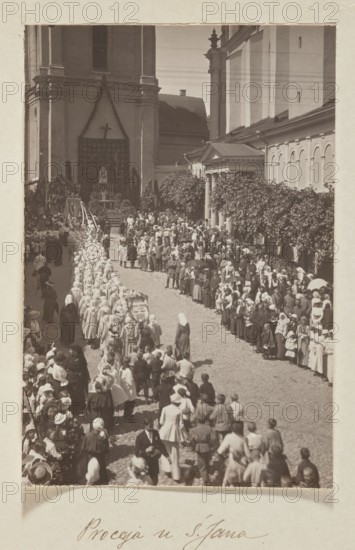 Procession at St. John's Church; photograph, Bulhak, Jan 1876–1950, St. John's Church Vilnius, 1916; photograph, photographic print: height 16.5 cm, width 11.2 cm; album page: height 31.0 cm, width 23.5 cm., The image shows a sepia-toned street scene with a procession moving toward a large building that appears to be a church; a group of people wearing light-colored head coverings and robes form a long line on a cobblestone street while crowds of onlookers stand along both sides of the street. The church facade and adjacent buildings are visible in shades of brown and beige, with architectural columns and a decorative doorway flanked by greenery; there is a raised platform or altar draped with fabric and a cross above it near the building entrance. Many individuals in the crowd wear hats, shawls, and coats in darker and lighter sepia tones, and a few people carry parasols; trees or climbing plants with darker foliage line the right side of the street. The overall palette is monochromat...