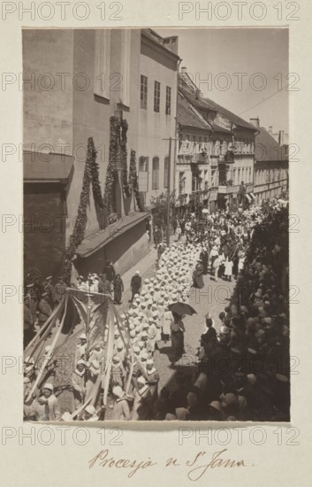 Procession at St. John's Church; photograph, 1916, Vilnius; subject: processions at St. John's Church Vilnius; maker: Jan Bulhak 1876–1950; medium: photographic print; dimensions: photograph 16.0 × 10.8 cm; album page 31.0 × 23.5 cm., A sepia-toned photograph showing a narrow cobblestone street crowded with a large procession of people, many wearing light-colored garments and hats, moving between tall buildings; a group near the front carries long flowing banners and ribbons, while other participants hold parasols and some wear darker clothing; buildings on the left display vertical greenery and windows, and balconies and awnings line the buildings on the right; a dense crowd of onlookers fills the sidewalks and casts shadows, and the overall palette is various shades of brown, beige, cream, and darker sepia tones.