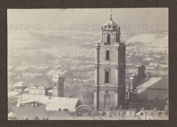 St. John's Church; photograph, 1914; subject: panoramic view of Vilnius including St. John's Church; photographer: Jan Bulhak 1876–1950; place of origin: Vilnius, Lithuania; medium: photographic print; image dimensions: 11.2 × 16.0 cm; album page dimensions: 31.0 × 23.5 cm., A grayscale photograph showing a tall, multi-tiered tower with arched openings and architectural detailing rising above a townscape; the tower is positioned toward the right side of the frame and is surrounded by a group of lower rooftops and building forms with sloped and flat surfaces. The foreground includes a cluster of roofs and chimneys, while the midground shows additional buildings and another smaller tower partially obscured. The background fades into distant, hilly terrain and soft atmospheric haze. Visible colors are shades of gray, off-white, and black.