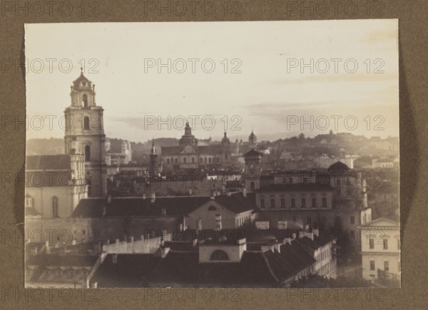 St. John's Church; photograph, 1912; panoramic view of Vilnius; photographer Jan Bulhak 1876–1950; photographic print, 11.5 × 16.2 cm; album page 31.0 × 23.5 cm., A sepia-toned cityscape photo mounted on a brown background showing a skyline of older buildings and church towers. The foreground has dark rooflines and lighter walls of several connected buildings with varied roof shapes. A tall multi-tiered bell tower stands on the left with architectural details and arched openings. In the midground there are more classical buildings and towers with domes and spires, and a larger building with a flat facade and evenly spaced windows. The background shows a hazy horizon with low hills and scattered tree silhouettes under a pale sky. Visible colors include brown, beige, cream, tan, and dark gray.
