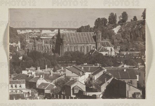 Bernardine Church; photograph, 1912, panoramic view of Vilnius; photographic print, height 10.5 cm, width 15.8 cm; album page height 31.0 cm, width 23.5 cm., The image shows a sepia-toned scene of a town with a group of rooftops in the foreground, composed of pitched roofs, chimneys, and brick and plaster facades in tones of brown and tan; behind the rooftops is a cluster of trees in darker browns and muted greens, and beyond the trees stands a large stone building with a steep roof and tall towers in darker brown tones; further back are rolling wooded hills with a mix of darker and lighter greenish-brown foliage and a lighter patch on a slope, and the sky above is a pale beige-gray.