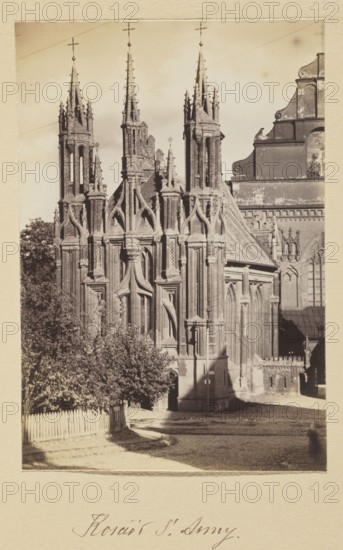 St. Anne's Church; photograph, 1912. Photographer: Jan Bulhak 1876–1950. Gothic style; St. Anne's Church Vilnius. Photographic print: 16.4 × 11.2 cm; album page: 31.0 × 23.5 cm., A sepia-toned photograph of a Gothic-style building with ornate vertical spires and pointed arches, showing intricate stone tracery and tall slender towers topped with finials. The facade displays layered buttresses and decorative window openings, with a steep roof section visible to one side. In front of the building there is a group of trees and a pale wooden picket fence, casting shadows on a light-colored ground surface. The image has a narrow light border around it and overall tones of brown, beige, and cream.