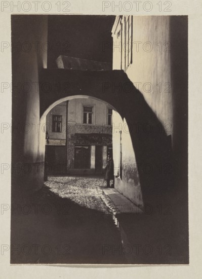 Bernardine Alley at night; photograph, ca. 1914, Jan Bulhak 1876–1950; Vilnius, Bernardine Alley; photographic print: 16.2 × 11.6 cm, album page: 31.0 × 23.5 cm., A dim archway frames a narrow cobblestone alley leading to the facade of an older building; a person stands beneath the arch near the right wall wearing a long dark coat and hat, partially in shadow. The scene shows strong contrasts of light and dark with deep black shadows under the arch and along the foreground, lighter cream and beige tones on the building walls, and muted gray and brown textures on the cobblestones and plaster. The arch casts a curved shadow across the ground and the right wall, and windows and architectural details are visible on the distant building.