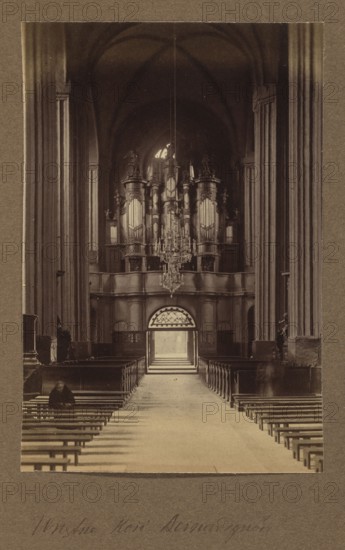 Interior of the Bernardine Church; photograph, ca. 1914; photographer Jan Bulhak 1876–1950; Vilnius, Bernardine Church; photographic print: height 16.0 cm, width 10.8 cm; album page: height 31.0 cm, width 23.5 cm., A tall interior view of a large vaulted space with a long central aisle flanked by wooden benches, a group of benches in the foreground showing their horizontal slats in shades of brown, a few figures seated or standing near the benches in darker tones, very tall columns rising on either side in beige and brown hues, an elevated ornate structure at the far end with decorative vertical elements and sculptural details in darker brown and muted gold tones, a large crystal chandelier hanging above the aisle with reflective highlights, an arched opening at the back letting in bright light that creates a pale rectangle at the end of the aisle, and an overall palette of sepia, brown, beige, and muted gold.