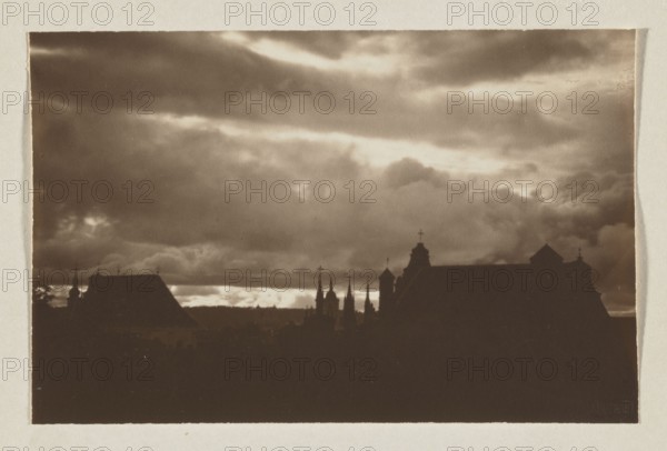 Churches and clouds; photograph, Jan Bulhak 1876–1950, Vilnius, 1912; panorama of Vilnius; photographic print, 10.3 × 15.9 cm; album page 31.0 × 23.5 cm., The image shows a dark sepia-toned scene with a cloudy sky occupying the upper portion and a silhouetted skyline of rooftops and spires along the lower portion; the clouds range from light tan to darker brown, and the silhouetted buildings appear as deep brown to near-black shapes with a few pointed spires and crosses visible against the lighter horizon.