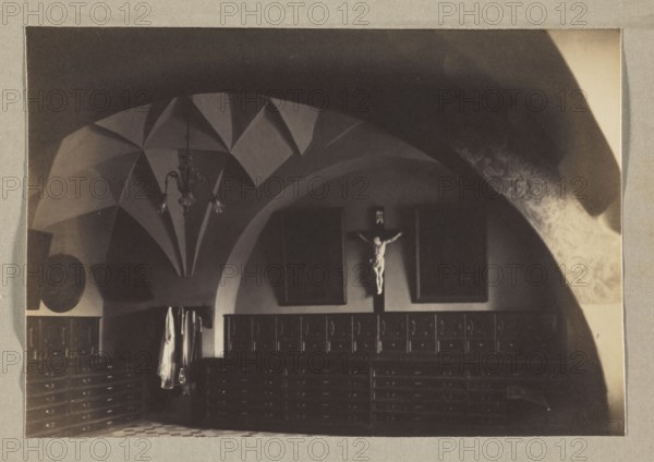 Sacristy at the Bernardine Monastery; photograph, ca. 1914; Bulhak, Jan 1876–1950; Bernardine Church, Vilnius; photographic print: height 11.3 cm, width 16.3 cm; album page: height 31.0 cm, width 23.5 cm., The image shows an interior room with vaulted ceilings and curved arches, featuring a hanging chandelier and decorative ceiling patterns. Along the walls are rows of wooden cabinets or drawers with paneled fronts and a group of hanging garments visible in one area. A crucifix figure is mounted on the wall between two dark framed panels. The floor has a patterned tile, and the visible colors in the image include shades of brown, beige, black, and cream.