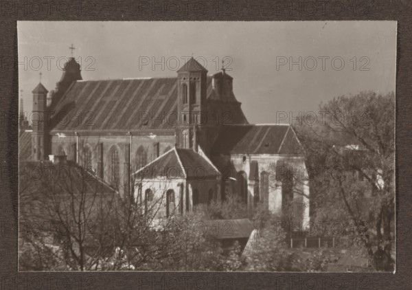 Bernardine Church; photograph, 1912; photographer Jan Bulhak 1876–1950; Vilnius; photographic print, 10.7 × 16.1 cm; album page 31.0 × 23.5 cm., A sepia-toned photograph shows a large stone building with steep pitched roofs and several towers, including a central tower with a pyramidal top and a smaller ornate tower with a curved roof; the building features tall arched windows and buttresses. In front of the main structure there is a smaller gabled annex with arched windows and a sloped roof. Leafless and leafy trees surround the lower part of the scene, partially obscuring some roofs and walls. The sky is plain and light in tone, and the image is framed by a dark textured border. Visible colors include shades of brown, tan, and muted gray.