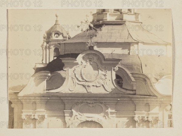 Dominican Church of the Holy Spirit Vilnius; photograph, 1912; Baroque architecture with domes; photographic print: 11.5 x 15.6 cm; album page: 31.0 x 23.5 cm., The image shows an ornate architectural facade with decorative curved moldings and sculpted embellishments in shades of beige and cream, with areas of darker brown and muted gray on the roof and shadows; a domed rooftop with a small ornamental finial and a taller tower with arched openings appear in the background in similar sepia tones; the overall palette is warm neutrals with soft contrasts between light and shadow.