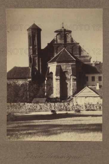 Bernardine Church; photograph, 1912; Bulhak, Jan 1876–1950; Vilnius; photographic print: height 16.0 cm, width 11.5 cm; album page: height 31.0 cm, width 23.5 cm, A sepia-toned photograph showing a large old stone building with architectural features including a tall tower on the left and a multi-tiered roof structure in the center, flanked by lower pitched roofs and a small building with a triangular gable on the right; the foreground contains a grassy area with lighter and darker bands of shadow, a low fence or wall, and a group of shrubs or low plants along the base of the buildings; visible colors include shades of brown, beige, tan, cream, and dark gray.