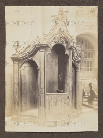 Confessional in the Dominican Church; photograph, Bulhak, Jan 1876–1950; Dominican Church of the Holy Spirit Vilnius; confessionals; Baroque; ca. 1914; photograph, photographic print: height 16.0 cm, width 11.9 cm; album page: height 31.0 cm, width 23.5 cm, The image shows an ornate wooden confessional booth with carved decorative scrollwork and curved arches, finished in warm brown tones; the interior appears darker brown and shadowed. The booth sits on a pale floor and is set against a background wall and large window area rendered in light beige and cream tones. In the background to the right there are several church pews with carved ends in dark brown, and the overall photograph has a sepia tint with soft contrasts.