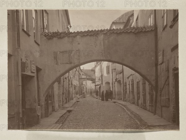 Jewish Street; photograph, album Wilno, volume 1, Jan Bulhak 1876–1950, Vilnius Jewish quarter, 1912; photograph, photographic print: 11.0 × 15.5 cm; album page: 31.0 × 23.5 cm., The image shows a narrow cobblestone street framed by an arched stone passage connecting two buildings, with a group of people standing under the arch further down the street. Buildings line both sides of the street with visible doorways and windows, some with shutters and worn facades. The street surface is uneven and bordered by raised sidewalks; another archway is visible farther along the street. The overall colors visible are sepia tones, brown, beige, and muted tan.