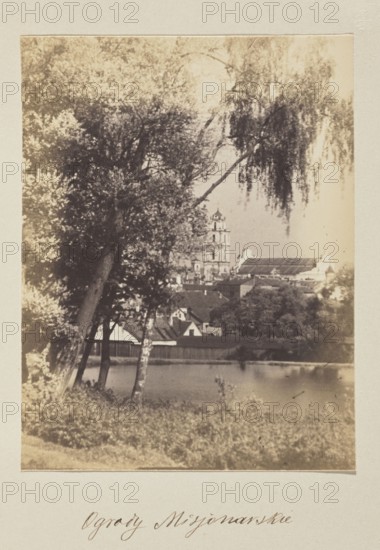 Missionary Gardens; photograph, 1912; Bulhak, Jan 1876–1950; missionary church and monastery, Vilnius; photographic print, 15.0 × 11.5 cm; album page 31.0 × 23.5 cm., A sepia-toned photograph showing a riverside scene with a group of tall trees in the foreground, including trees with dark trunks and hanging branches and lighter foliage; a body of water is visible behind the trees with a reflective surface. Beyond the water there are several buildings with pitched roofs and a larger multi-tiered tower rising above them. The image contains areas of light beige, brown, dark brown, and muted gray tones throughout.