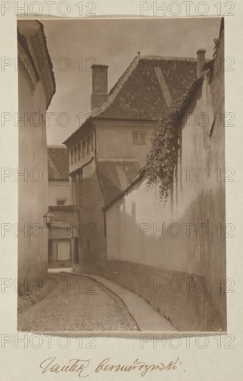 Bernardine Alley; photograph, ca. 1912, Jan Bulhak 1876–1950; Vilnius Lithuania, Bernardine Alley; photographic print: 16.2 × 10.8 cm; album page: 31.0 × 23.5 cm, A narrow cobblestone alley bordered by tall walls and buildings in tones of brown and beige, with a smooth light-colored sidewalk along one side; a tiled roof with darker brown and tan tiles slopes above, showing a chimney and a small window; a hanging lantern projects from a wall near an arched passageway that leads deeper into the alley; a vine with darker foliage cascades down one wall, casting soft shadows across the lighter wall surfaces; the sky above is a muted brownish-gray.