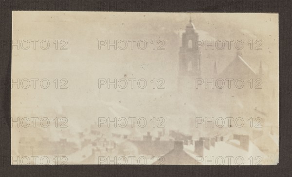 All Saints' Church; photograph, 1912, panorama of Vilnius; photographer Jan Bulhak 1876–1950. Photographic print: 9.5 × 16.5 cm; album page: 31.0 × 23.5 cm., A faded, sepia-toned scene showing a hazy cityscape with rooftops in the foreground and a tall, tower-like structure with a peaked roof and spires faintly visible in the background; the sky and buildings blend into pale beige and light brown tones, with several darker brown smudges and spots scattered across the image and a dark brown border framing the picture.