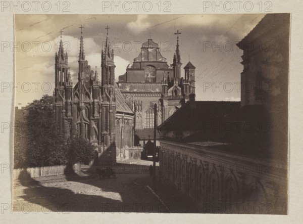 Church of St. Anne and the Bernardines; photograph, 1912. Subject: Bernardine Church Vilnius and St. Anne's Church Vilnius, Gothic style. Photographer: Jan Bulhak 1876–1950. Medium: photographic print; image size 11.6 x 15.8 cm; album page size 31.0 x 23.5 cm., A sepia-toned photograph showing a street flanked by architectural structures, with a large ornate Gothic-style church dominating the left and center featuring tall spires, pointed arches, and intricate stonework, and a long building with repeating arched details running along the right side and foreground. A few trees and foliage appear near the church, and a low picket fence lines part of the street. Shadows stretch across a cobbled or textured roadway where a small cart or wagon and a couple of indistinct figures are visible. The sky shows a few clouds above the buildings. The visible colors are shades of brown, tan, beige, and muted gray.