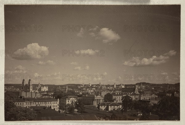 Vue générale Vilnius. General View; photograph, pictorialism, panorama of Vilnius, ca. 1913; photograph, photographic print: height 10.8 cm, width 16.4 cm; album page: height 31.0 cm, width 23.5 cm., A wide, sepia-toned view of a city skyline beneath a sky filled with a few fluffy clouds, showing a variety of buildings with classical and baroque architectural features, including several church towers with spires and domes, a prominent building with a columned façade, rows of multi-story structures with pitched roofs, and groups of trees interspersed among the buildings; the overall palette includes brown, beige, tan, cream, and darker brown tones.