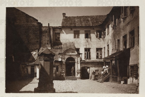 Vilnius — Ghetto. Yard in the Jewish Quarter. Photographer: Jan Bulhak 1876–1950. 1914. Photograph print glued to light-toned cardboard. Subject: courtyards, tenement houses in Vilnius, Lithuania. Photographic print: 11.5 × 16.7 cm; cardboard: 31.9 × 24.1 cm., The image shows an old cobblestone courtyard surrounded by multi-story buildings with tiled roofs and many windows, rendered in sepia tones of brown, beige, and dark tan; a central ornate wooden pillar or well structure with a small peaked roof stands in the foreground casting a shadow, while a doorway with an arched pediment and steps sits centrally at the far side with a person standing in the entrance; to the right a wooden staircase leads up to a landing where a few people are gathered, and there are additional doors and windows along the surrounding façades with varied textures of plaster and brick.