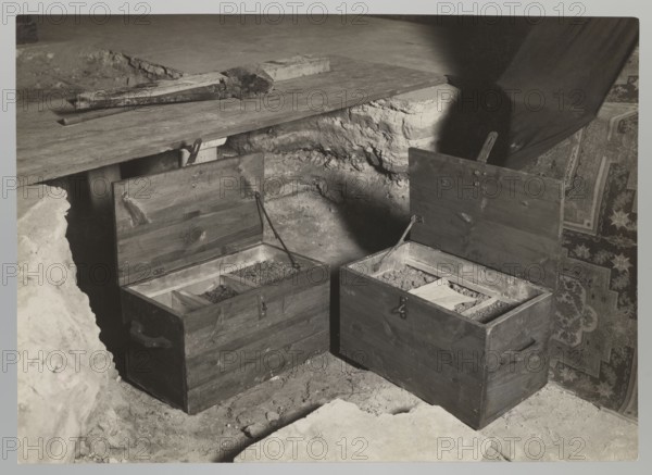 Royal Cathedral Crypt — Open Chests; Bulhak, Jan 1876–1950; Vilnius Lithuania; Archcathedral Basilica of St. Stanislaus and St. Ladislaus; crypts; 1931 flood in Vilnius; 1931; photograph; nature photograph; height 12.0 cm; width 16.0 cm, The image shows a grayscale scene of an indoor area with a rough stone or dirt floor and a wooden platform; two wooden chests with open lids sit side by side on the floor, each chest divided into compartments containing loose granular material; the chests have dark wood tones and metal hinges and handles, and there are a few tools or wooden pieces resting on the platform above; a patterned textile with intricate designs hangs or is draped on the right side, showing darker and lighter areas; shadows fall across the scene, creating contrasts between light and dark gray tones.