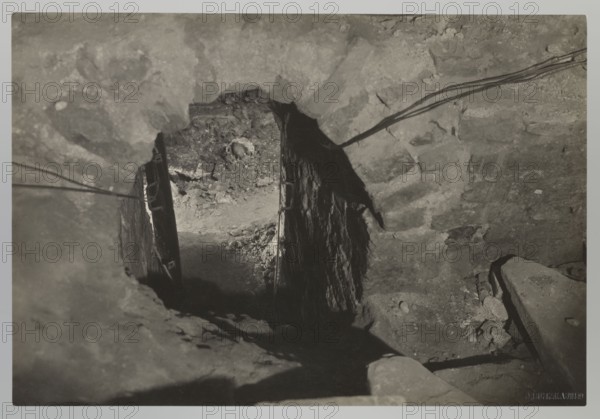 Vilnius. Cathedral, entrance to the royal crypt, Bulhak, Jan 1876–1950, Vilnius Lithuania, Archcathedral Basilica of St. Stanislaus and St. Ladislaus, crypts, archaeological excavations, flood in Vilnius 1931, 1931, Vilnius, photograph from nature, height 11.5 cm, width 16.5 cm, The image shows a dark, rocky interior space with rough stone surfaces and a jagged opening leading down into a shadowed area; a group of cables or wires run along the upper right across the stone, and a rough wooden beam or plank is visible near the opening with metal fittings; the ground inside the opening is uneven with small rocks and debris, and there are areas of deep shadow and lighter stone texture creating contrast in tones of gray, black, and off-white.