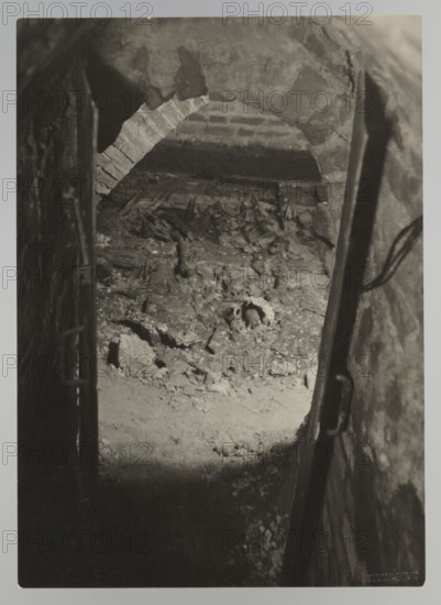 Vilnius. Cathedral — entrance to the royal crypt; photograph by Jan Bulhak 1876–1950; Archcathedral Basilica of St. Stanislaus and St. Ladislaus, Vilnius; crypts; archaeological excavations after the 1931 flood in Vilnius; 1931; photograph from nature; 11.5 × 16.5 cm., The image shows a dim, narrow underground passage with a rough arched brick opening leading into a low chamber filled with rubble and uneven earth; the scene contains a group of broken and jagged fragments scattered on the floor and a partially buried round object among the debris, with shadowed walls flanking the passage and a dark vertical element on the right that appears to be a metal bar or support; visible colors are shades of gray, black, and off-white.