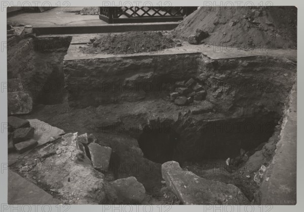 Vilnius. Cathedral - entrance to the royal crypt; Bulhak, Jan 1876–1950; Vilnius Lithuania; Archcathedral Basilica of St. Stanislaus and St. Ladislaus Vilnius; crypts; archaeological excavations; flood in Vilnius 1931; 1931; Vilnius type of place / place of origin; photograph; natural photography; height 11.5 cm; width 16.7 cm, The image shows a excavation pit with exposed earth layers and rough stone edges, surrounded by broken slabs and scattered rubble; a group of piled stones sits on a ledge above the pit, and a dark circular opening is visible within the exposed soil; the scene includes a raised flat surface at the top with a lattice fence section and a mound of loose earth behind it; visible colors are shades of gray, black, and white.