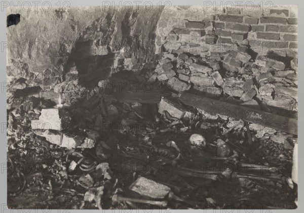 Vilnius. Cathedral — interior of the Kiezgajlowska Crypt. Photographer: Jan Bulhak 1876–1950. Archcathedral Basilica of St. Stanislaus and St. Ladislaus, Vilnius. Crypts; archaeological excavations; graves; flood in Vilnius, 1931. Photograph; nature photograph. Dimensions: height 12.0 cm, width 17.0 cm., The image shows an indoor scene with a rough stone and brick wall and a floor cluttered with rubble and scattered bones, including a visible skull among the debris. A few broken bricks and larger stone slabs lie across the floor, and a small candle sits atop a stack of bricks casting light. The visible colors are shades of black, white, and gray.