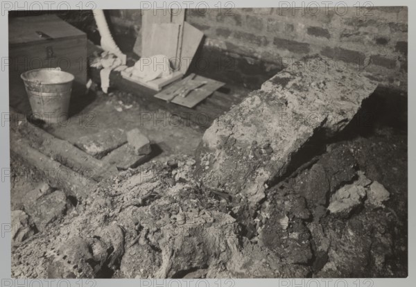 Vilnius. Cathedral — interior of the royal crypt. Photograph by Jan Bulhak 1876–1950, 1931. Depicts crypts revealed by archaeological excavations after the 1931 flood in Vilnius. Photograph from nature; 14.5 × 16.5 cm., A grayscale photograph showing a rough indoor space with a brick wall backdrop and a dirt floor scattered with broken concrete and rubble; a large irregular slab of concrete or stone leans across the right side, and a pile of crumbled material with coarse texture occupies the foreground. Toward the left a metal bucket sits near a wooden chest and a group of wooden boards and planks, with a few pieces of cloth draped over them. Several rectangular blocks and pieces of masonry lie on the ground around a shallow trench or depression. The visible tones are various shades of gray, black, and white throughout the scene.