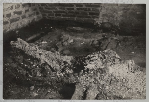 Vilnius — Cathedral interior of the royal crypt. Photograph by Jan Bulhak 1876–1950. Archcathedral Basilica of St. Stanislaus and St. Ladislaus Vilnius, crypts; archaeological excavations; tomb of Barbara Radziwillówna Queen of Poland, 1520–1551. Flood in Vilnius, 1931. Photograph from nature; 1931. Height 11.5 cm; width 17.0 cm., The image shows a dimly lit interior corner with a rough brick wall in the background and a dirt floor in the foreground, featuring a large, irregular mound of crumbled material and debris spread across the ground; the scene includes textured, fragmented objects partially buried in the dirt and a few scattered pieces of rubble, with visible tones of black, white, gray, and varying shades of sepia.