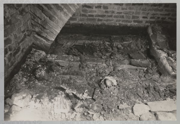Vilnius. Cathedral — interior of the royal crypt. Photograph from nature, 1931. Depicts crypts and archaeological excavations following the 1931 flood in Vilnius. Photographer: Jan Bulhak 1876–1950. Place: Vilnius Lithuania, Archcathedral Basilica of St. Stanislaus and St. Ladislaus. Photograph; 11.5 × 16.5 cm., The image shows a dim interior space with brick walls and an arched brick structure, a rough dirt and debris-covered floor, and scattered wooden beams and broken planks. There are several irregular chunks of rubble and fragments of material spread across the ground, along with a partially exposed rounded object among the debris. The visible colors are shades of gray, black, and white.