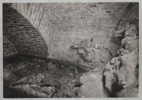 Cathedral interior of the royal crypt, Bulhak, Jan 1876–1950, Vilnius, Archcathedral Basilica of St. Stanislaus and St. Ladislaus, crypts, archaeological excavations, flood in Vilnius 1931, 1931, photograph from nature, height 12.0 cm, width 17.0 cm, A dim interior scene showing a rough brick arch and brick walls meeting a stone floor strewn with debris, featuring a sloped brick arch on the left, a textured brick wall at the back, and irregular stone and rubble formations on the right and foreground; visible materials include brick, stone, dirt, and loose fragments, and the image appears in shades of gray, black, and white.