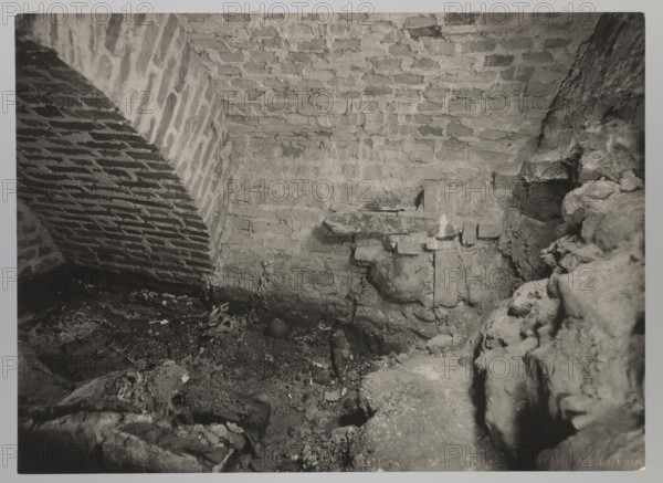 Vilnius. Cathedral — interior of the royal crypt; photograph by Jan Bulhak 1876–1950, 1931. Subject: Archcathedral Basilica of St. Stanislaus and St. Ladislaus, royal crypts; archaeological excavations after the 1931 flood in Vilnius. Natural photograph; 12.0 × 16.0 cm., The image shows an interior scene of a rough brick and stone space with a vaulted brick arch on the left and a brick wall and ceiling occupying the center and upper area; the floor area is uneven and covered with dirt, rubble, and broken fragments, and there are irregular stone formations and mortar along the right side. The visible colors are shades of gray, black, and off-white.