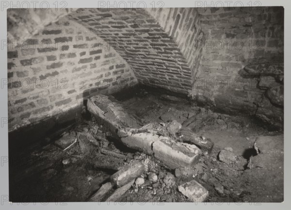 Vilnius. Cathedral — interior of the royal crypt, photographed by Jan Bulhak 1876–1950. Archcathedral Basilica of St. Stanislaus and St. Ladislaus, crypts; archaeological excavations; associated with Barbara Radziwillówna Queen of Poland, 1520–1551. Flood in Vilnius, 1931. Photograph from nature; gelatin silver print, — height 11.5 cm, width 16.5 cm., The image shows a dimly lit interior space with brick walls and a curved brick ceiling; the bricks display various shades of gray. In the foreground there is a damaged, broken rectangular stone or concrete object surrounded by rubble and fragments, all in tones of gray and black. The floor appears uneven and dirty with scattered chunks of masonry and loose debris in darker and lighter grays. Shadows create contrast along the base of the walls and among the rubble, producing areas of deep black and mid-gray. Overall the visible colors are shades of gray, black, and white.