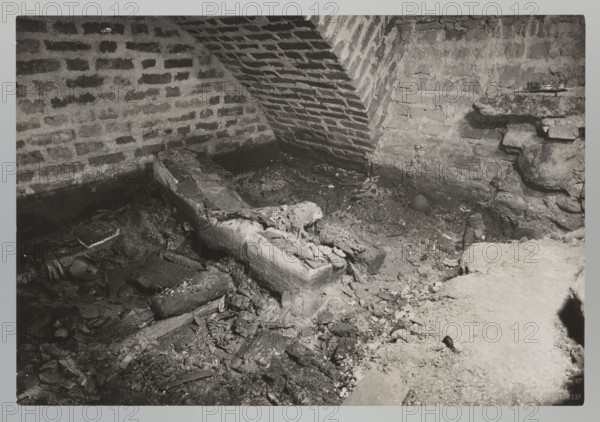 Vilnius. Cathedral — interior of the royal crypt, Bulhak, Jan 1876–1950, Vilnius Lithuania, Archcathedral Basilica of St. Stanislaus and St. Ladislaus, crypts, archaeological excavations, flood in Vilnius in 1931, 1931, photograph, natural photography, height 11.5 cm, width 17.0 cm, A dim, grayscale scene of a rough brick-walled corner with an arched brick support, a rubble-strewn floor, and a large broken, decayed wooden beam or log lying diagonally; the walls and floor show crumbling mortar and debris, with scattered fragments of wood and plaster, a small round object partially buried, and areas of darker and lighter tones creating contrast between the shadowed left side and the brighter right foreground.