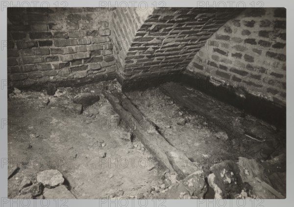 Vilnius. Cathedral — interior of the royal crypt, photograph by Jan Bulhak 1876–1950, Archcathedral Basilica of St. Stanislaus and St. Ladislaus, crypts, archaeological excavations after the 1931 flood in Vilnius, 1931, photograph from nature, 11.5 × 16.5 cm, The image shows an interior corner with brick walls meeting a sloped brick ceiling and a rough dirt floor scattered with debris and fragments of material; a few long, dark wooden beams lie across the floor and a group of stones or rubble sits near the foreground; visible colors include various shades of gray, black, and off-white.