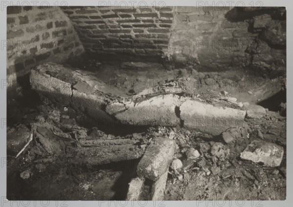 Vilnius. Cathedral — interior of the royal crypt, photographed by Jan Bulhak 1876–1950, Archcathedral Basilica of St. Stanislaus and St. Ladislaus Vilnius, crypts, archaeological excavations after the 1931 flood in Vilnius, 1931. Photograph; natural photography. Image size: 11.5 cm × 16.7 cm., The image shows a dim interior scene with a rough brick wall in the background and a broken, curved stone or concrete structure lying among rubble in the foreground. The surface textures include cracked and crumbling masonry, loose debris, and scattered fragments of rock and mortar. Lighting is low and creates strong contrasts and shadows across the uneven surfaces. Visible colors are shades of gray, black, and off-white.